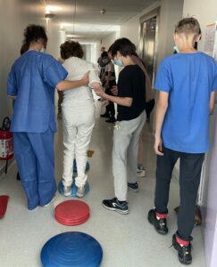 Un étudiant et une professionnelle de santé aide un personne agée à se déplacer pour traverser un parcours proprioceptif, organisé dans le couloir de l'hôpital.